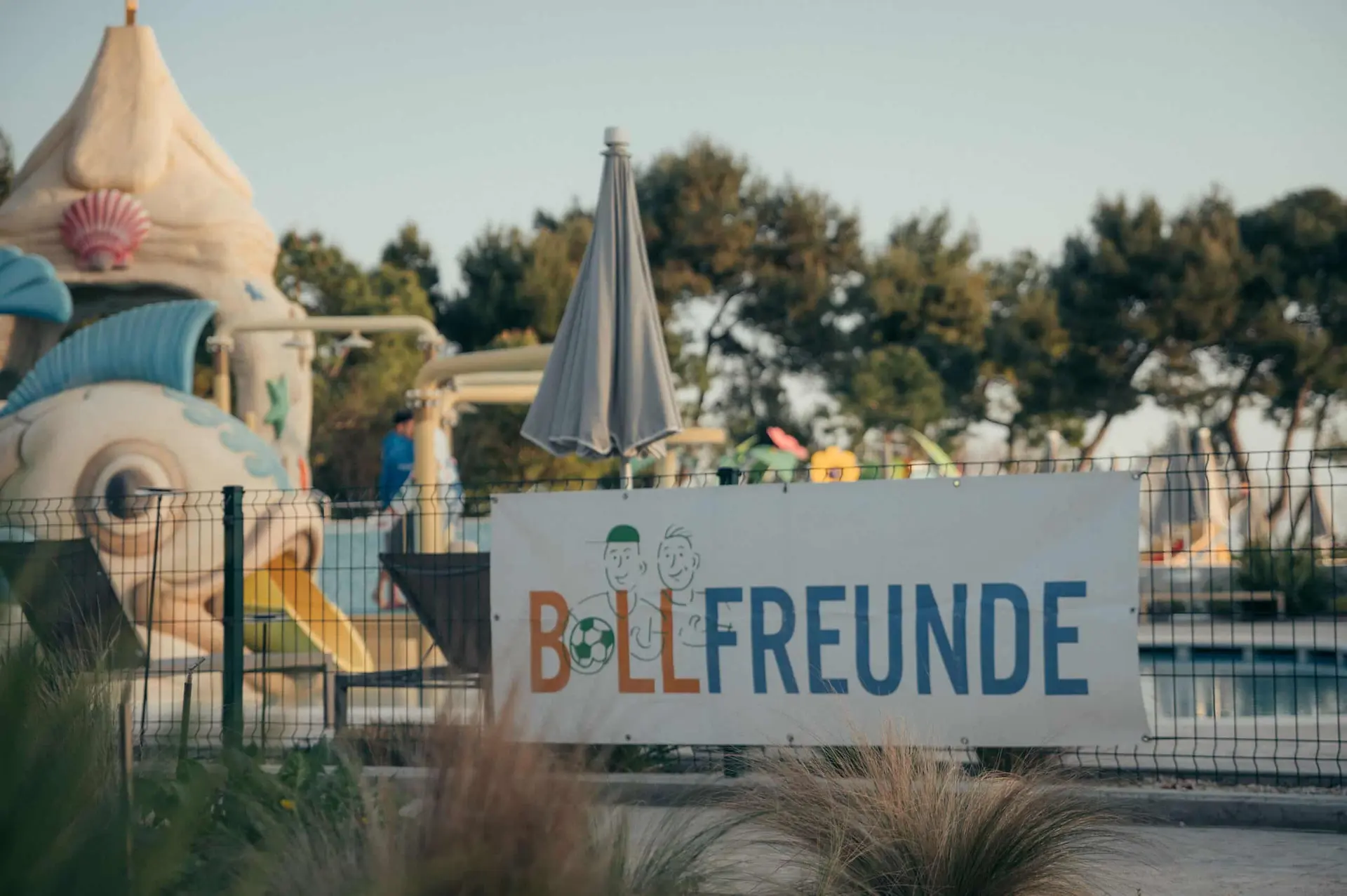 BALLFREUNDE Schild an Zaun vor einem Pool mit Rutsche und Liegen